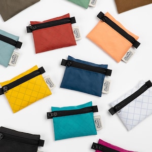 Könnte beinhalten: Eine Sammlung kleiner, farbenfroher Reißverschlusstaschen in verschiedenen Farbtönen, darunter Rot, Gelb und Blau. Jede Tasche hat einen schwarzen Reißverschluss und ein kleines weißes Etikett mit schwarzem Text. Die Taschen sind auf einer weißen Oberfläche angeordnet.