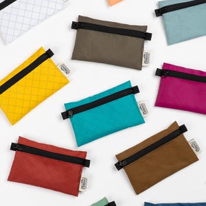 Könnte beinhalten: Eine Sammlung kleiner, rechteckiger Beutel in verschiedenen Farben, darunter Gelb, Türkis und Braun. Jeder Beutel hat einen schwarzen Reißverschluss und ein kleines Stoffetikett. Die Beutel haben ein Rautenmuster.