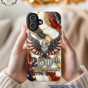 Peut inclure: Une coque de téléphone avec un fond en marbre blanc et des accents rouges, bleus et dorés. Un aigle à tête blanche aux ailes déployées est représenté au-dessus du texte 250th ANNIVERSARY 1776-2026. L'aigle et le texte ont un aspect brillant et diamanté.