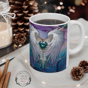 Op de afbeelding: Witte keramische mok met een ontwerp van witte engelen vleugels en een turquoise hartvormige edelsteen. De mok heeft een handvat en is gevuld met een donkere vloeistof. De mok is versierd met gouden accenten en de woorden "Sip & Style" staan onderaan.