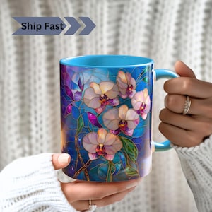 May include: A blue ceramic mug with a stained glass design featuring white orchids with purple accents. The mug has a handle and is filled with a light blue liquid.