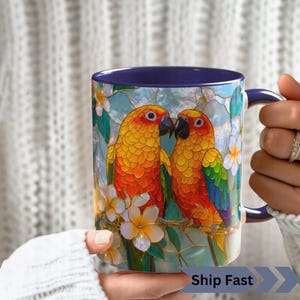 May include: A ceramic mug with a dark blue interior and handle, featuring a vibrant stained-glass style illustration of two sun conure parrots perched on a branch surrounded by white tropical flowers against a light blue sky background.
