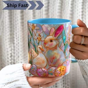 May include: A ceramic mug with a light blue interior and handle, featuring a colourful stained-glass style illustration of a rabbit, a duckling, and decorated Easter eggs surrounded by white lilies against a soft pastel background.