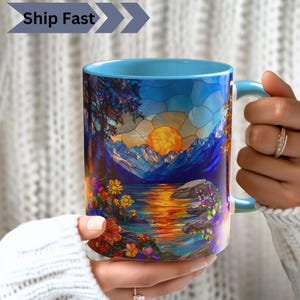 May include: A ceramic mug with a light blue handle and interior, featuring a vibrant stained-glass style print of a sunset over mountains, a lake, and colorful wildflowers. The mug displays a graphic design with deep blues, oranges, and yellows.
