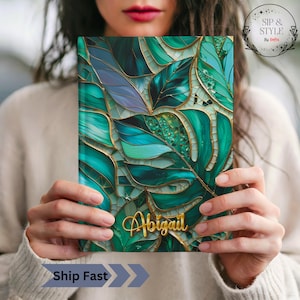 May include: A journal with a vibrant, abstract design of green and blue leaves with gold accents. The name "Abigail" is written in gold script. The journal is held by a person wearing a cream-colored sweater. The text "Ship Fast" is also visible.