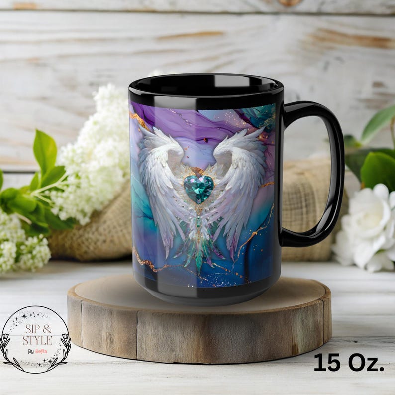 Op de afbeelding: Zwarte koffiemok van 444 ml met een ontwerp van witte engelen vleugels, een teal hart en een kleurrijke achtergrond. De mok heeft de tekst "Sip & Style" en "15 Oz." erop gedrukt.