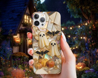 May include: A decorative phone case featuring a Halloween theme. The case has a cream-colored background with gold accents and includes images of ghosts, bats, and pumpkins. The case is held in a hand against a background of a spooky night scene.