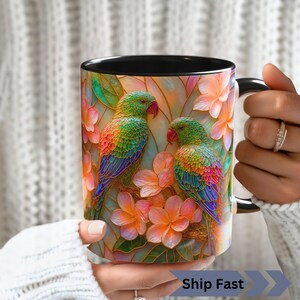 May include: A ceramic mug with a black interior and handle, featuring a vibrant, stained-glass style illustration of two green parrots perched among soft pink and orange tropical flowers with green leaves, set against a light background.