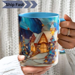 Puede incluir: Una taza de cerámica con interior y asa de color azul claro, que presenta una colorida ilustración estilo vidriera de un pueblo invernal con casas cubiertas de nieve, ventanas de color amarillo brillante y pinos decorados bajo un cielo azul oscuro.