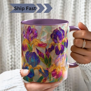 Può includere: Una tazza in ceramica con manico e bordo viola, caratterizzata da una stampa vivace in stile vetrata con fiori di iris viola, blu e oro su uno sfondo madreperlaceo. La tazza presenta un motivo floreale dettagliato e dai colori brillanti.