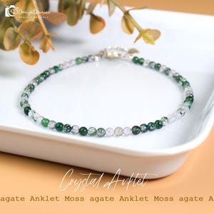 May include: A delicate anklet featuring small, round beads in shades of green and clear. The beads are strung together with a silver-colored chain. The text "Crystal Anklet" is visible in the image. The anklet is displayed on a white surface.