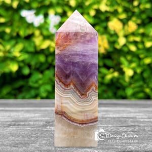 May include: A purple, white, and orange banded crystal point. The crystal is polished and has a pointed tip. The crystal is sitting on a wooden surface.