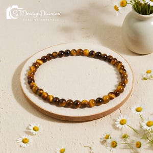 May include: A beaded bracelet made of tiger's eye gemstones. The bracelet is a warm brown color with golden and dark brown variations. The beads are strung together to form a circular shape. The bracelet is displayed on a round, light-colored surface.