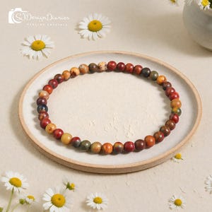May include: A bracelet made of multi-coloured beads, including red, brown, yellow, and green, sits on a white plate. The bracelet is surrounded by daisies. The image includes the text "SJ Design Diaries Healing Crystals."