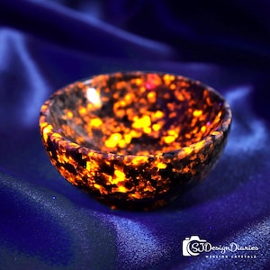 May include: A small, round bowl with a speckled pattern of orange and black. The bowl is made of a stone material and has a smooth, polished finish. The bowl is set against a dark blue background.