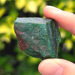 May include: A raw, unpolished mineral specimen featuring a deep forest green color with visible patches of dark brown and reddish-brown inclusions. The stone has a rough, textured surface with irregular edges and a dense, opaque appearance.