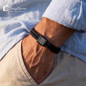May include: Black rope bracelet with a square clasp. The clasp features the text "26" and the letters "L", "E", and "O" arranged around a symbol. The bracelet is worn on a wrist, with a light blue shirt and tan pants in the background.