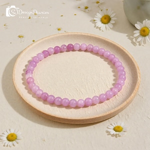 Könnte beinhalten: Ein kreisförmiges Perlenarmband bestehend aus kleinen, glatten und polierten Edelsteinkugeln in hellen Violett- und Lavendeltönen. Das Armband ist ringförmig angeordnet und zeigt eine einheitliche Größe sowie Textur bei jeder einzelnen Perle.