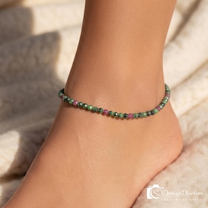 May include: Ankle bracelet featuring small, round beads in shades of green and pink. The beaded bracelet is worn around the ankle. The image is taken in natural light, highlighting the colors of the gemstones.