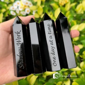 Può includere: Quattro obelischi di cristallo di ossidiana nera con punte. Due obelischi hanno incise le parole "Shadow Work" e "One day at a time". I cristalli sono tenuti in una mano, con un fogliame verde sullo sfondo.