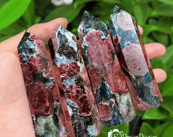 May include: Four polished crystal towers with a mix of red, green, and blue hues. The crystals are held in a hand against a backdrop of green foliage. The crystals have a pointed top and a rectangular base. The crystals are a natural stone.