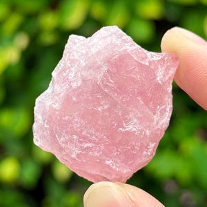 May include: A rough, pink rose quartz crystal with a natural, irregular shape. The crystal is translucent and has a light pink hue.