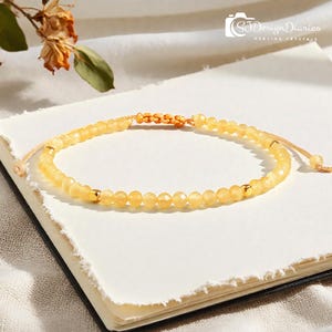 May include: A delicate yellow beaded bracelet with gold accents. The bracelet is made of small, faceted yellow beads strung together on a neutral-colored cord. The bracelet is resting on a white surface. The text "SJ Design Diaries Healing Crystals" is in the upper right corner.