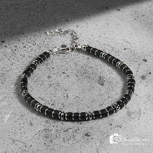 May include: A bracelet featuring alternating matte black cylindrical beads and polished silver-tone metal spacers. The piece includes a silver-tone metal chain extender and a lobster claw clasp, creating a uniform and repeating pattern throughout the design.