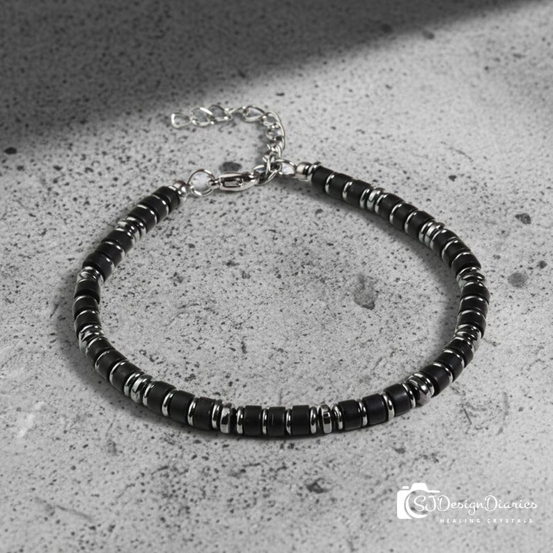 May include: A bracelet featuring alternating matte black cylindrical beads and polished silver-tone metal spacers. The piece includes a silver-tone metal chain extender and a lobster claw clasp, creating a uniform and repeating pattern throughout the design.