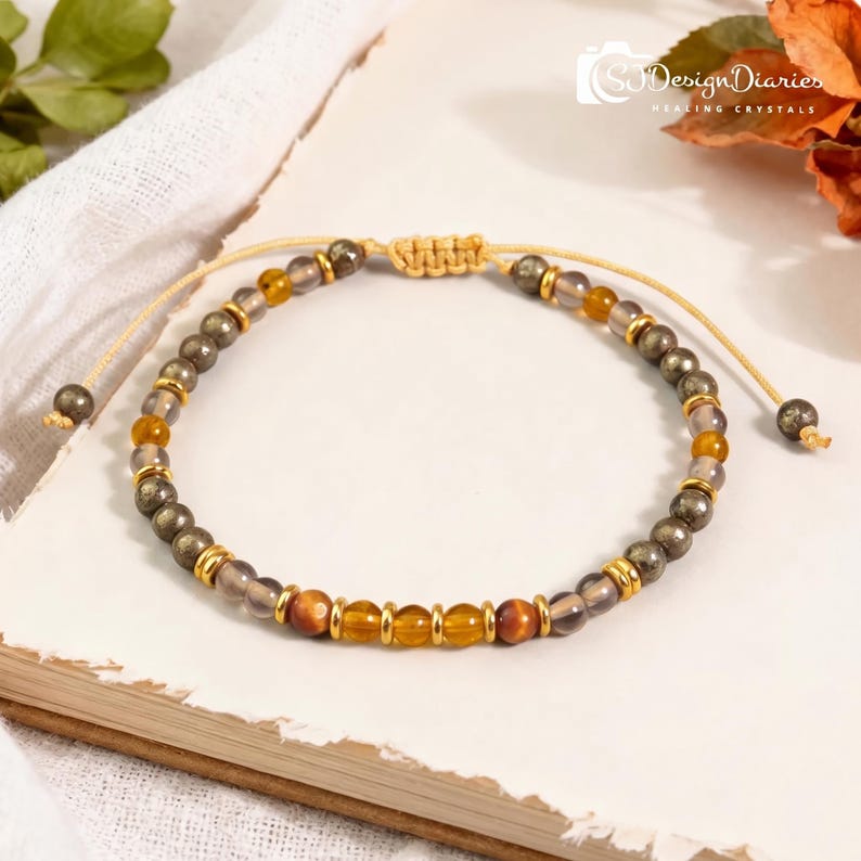 K&ouml;nnte beinhalten: Ein Perlenarmband mit abwechselnden dunkelgrauen metallischen Perlen, durchscheinenden bernsteinfarbenen Perlen und braunen Tigeraugen-Perlen, akzentuiert durch kleine goldfarbene Metallabstandshalter. Das Armband hat eine beigefarbene Kordel.
