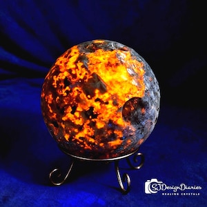 May include: A glowing, spherical decorative object with a fiery orange and black pattern, resting on a delicate, curved metal stand. The sphere has a textured surface and a dark blue background. The object resembles a celestial body.