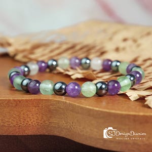 Könnte beinhalten: Ein Armband mit einer Mischung aus lila, grünen, klaren und dunkelgrauen Perlen. Das Armband besteht aus Natursteinen und wird auf einer Holzoberfläche präsentiert. Das Bild enthält den Text "SJDesignDiaries Healing Crystals".