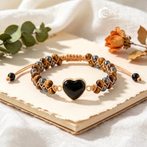 May include: A braided cord bracelet featuring alternating brown tiger eye beads and metallic gray hematite beads. The center of the bracelet showcases a smooth, black heart-shaped charm set in a gold-toned frame, with adjustable cord ends.