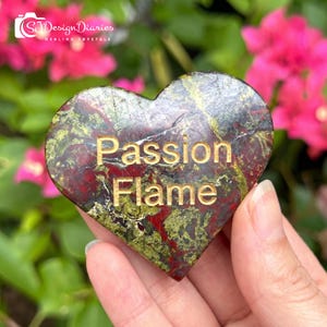 May include: A heart-shaped polished stone featuring a mottled pattern of deep red and olive green colours. The words Passion Flame are engraved in gold lettering across the centre of the stone surface, which has a smooth and glossy finish.