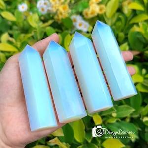 May include: Four iridescent white crystal points, also known as angel aura quartz, are held in a hand. The crystals are smooth and polished, with a slight blue hue.