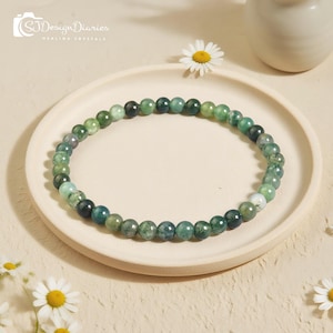 May include: A beaded bracelet made of green and dark green gemstones. The bracelet is displayed on a white plate with a daisy and the text "SJ Design Diaries Healing Crystals".