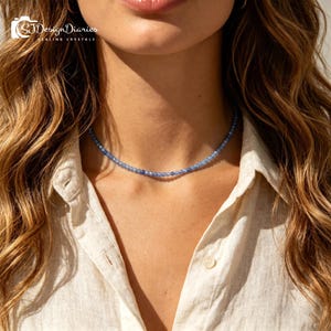 May include: A delicate choker necklace featuring small, round, faceted blue beads. The necklace sits closely around the neck, showcasing a uniform arrangement of translucent, sapphire-toned stones that catch the light with a subtle sparkle.