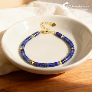 May include: A blue lapis lazuli bracelet with gold-colored accents and a heart-shaped charm. The bracelet is composed of small, square-shaped beads and is displayed in a white bowl. The clasp and connecting chain are gold-colored.