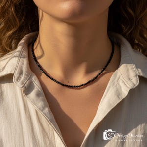 May include: A delicate necklace composed of small, faceted black beads worn around the neck. The beads are uniform in size and shape, creating a continuous, dark, reflective strand that rests against the skin. The necklace is worn with a light-colored shirt.