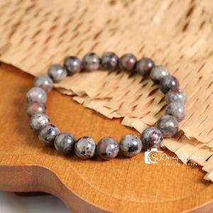 May include: A beaded bracelet featuring round, gray and maroon speckled stones. The bracelet is displayed on a wooden surface with a light brown paper background. The text "SJ Design Diaries Healing Crystal" is visible.