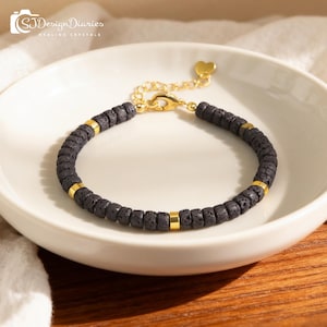 Puede incluir: Una pulsera de piedra de lava negra con detalles dorados. La pulsera presenta pequeñas cuentas oscuras separadas por espaciadores cilíndricos dorados. El cierre y un pequeño dije de corazón también son dorados. La pulsera se exhibe en un plato de cerámica blanco.