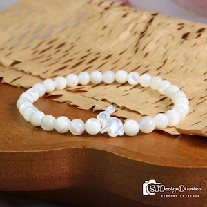 May include: White beaded bracelet featuring a small, pearlescent bunny charm. The bracelet is made of round, white beads and is displayed on a wooden surface. The bracelet is a piece of jewelry.