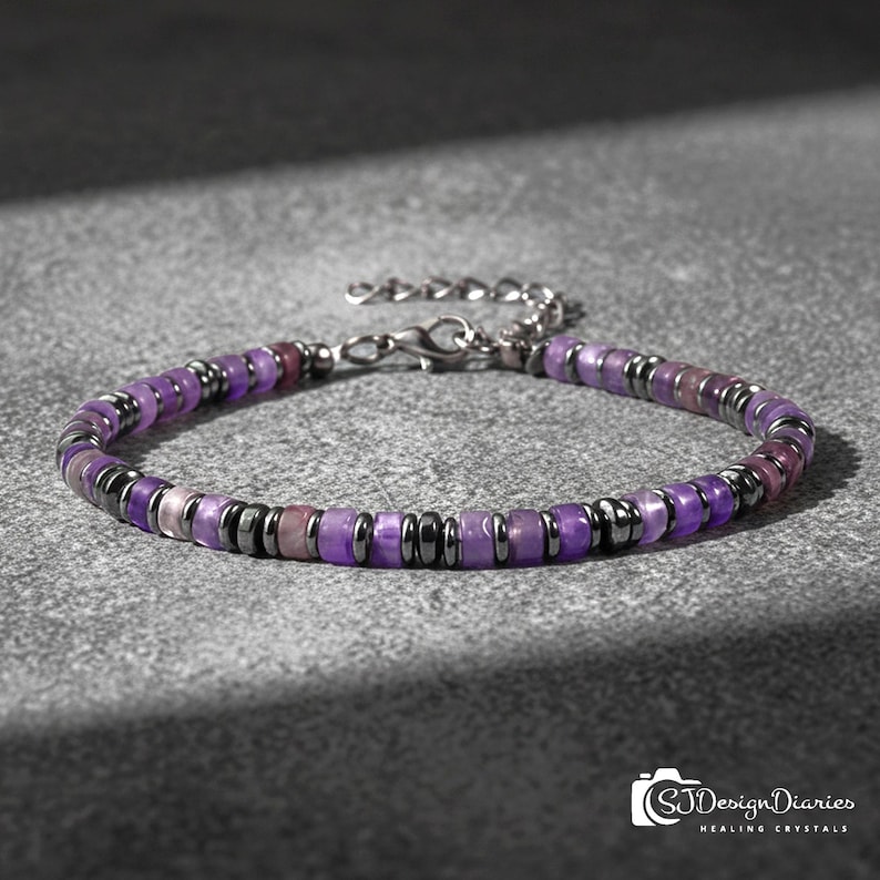 May include: A beaded bracelet featuring purple and gray beads. The bracelet has a silver-colored clasp and chain. The beads are a mix of purple and gray, with a few pinkish beads interspersed. The bracelet is on a gray surface.