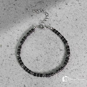 May include: A bracelet featuring a repeating pattern of small, matte black cylindrical beads separated by thin, polished silver-tone metal spacers. The piece includes a silver-tone lobster claw clasp and an adjustable chain link extension at the end.