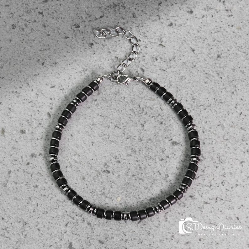 May include: A bracelet featuring a repeating pattern of small, matte black cylindrical beads separated by thin, polished silver-tone metal spacers. The piece includes a silver-tone lobster claw clasp and an adjustable chain link extension at the end.