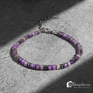 May include: A beaded bracelet featuring purple and pink gemstones and dark gray metal spacers. The bracelet has a silver-colored chain and clasp. The image is on a gray surface with a shadow.