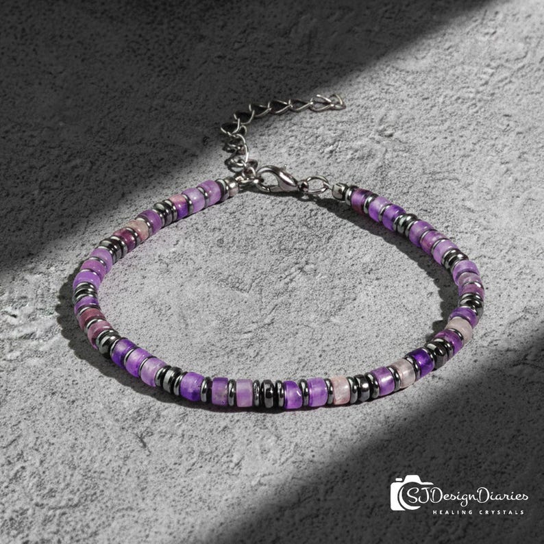 May include: A beaded bracelet featuring purple and pink gemstones and dark gray metal spacers. The bracelet has a silver-colored chain and clasp. The image is on a gray surface with a shadow.