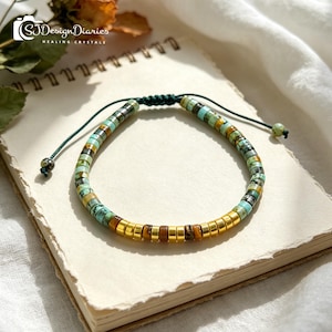 May include: A beaded bracelet with turquoise, gold, and brown accents. The bracelet is made of small, round beads strung together on a dark green cord. The bracelet is resting on a notebook.