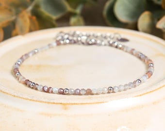 May include: Delicate beaded bracelet featuring small, multi-colored gemstones and silver-toned metal accents. The bracelet is displayed on a cream-colored plate, with green foliage in the background. The gemstones appear to be approximately 0.1 inches in diameter.