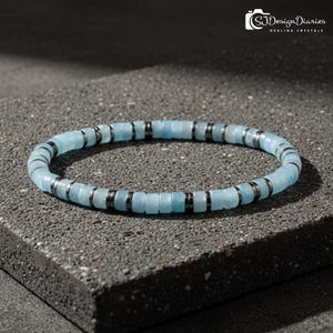 Op de afbeelding: Een lichtblauwe kralenarmband met donkergrijze metalen accenten. De armband is gemaakt van kleine, vierkant gesneden kralen en wordt getoond op een grijze ondergrond. De tekst "SJ Design Diaries Healing Crystals" is zichtbaar.